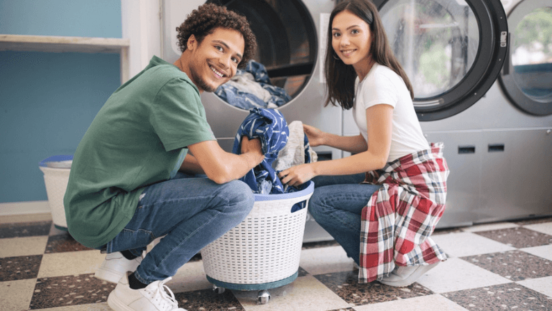 Happy Young Multicultural Couple Doing Laundry Together Loading Washer Machine At Laundromat Room.