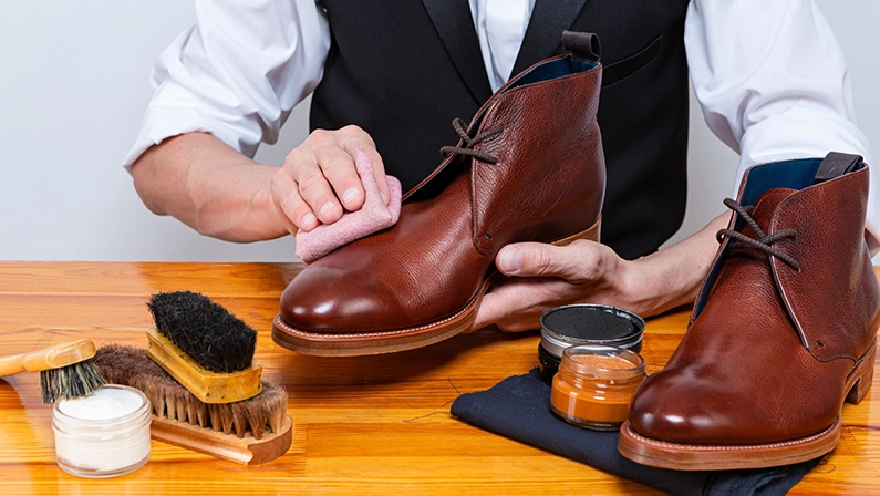 Professional Male Shoes Cleaner with Cleaning Brushes For Tan Derby Leather Boots While Working in Workshop With Rub Cloth. Horizontal Image