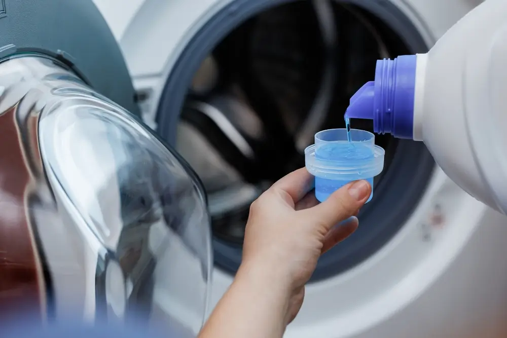 Woman pours liquid washing gel into plastic cap against background of drum of steel-colored washing machine.