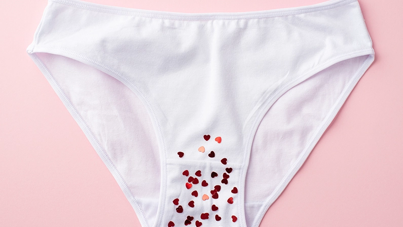 Top view photo of white cotton classic panties with red heart shaped confetti on isolated pastel pink background