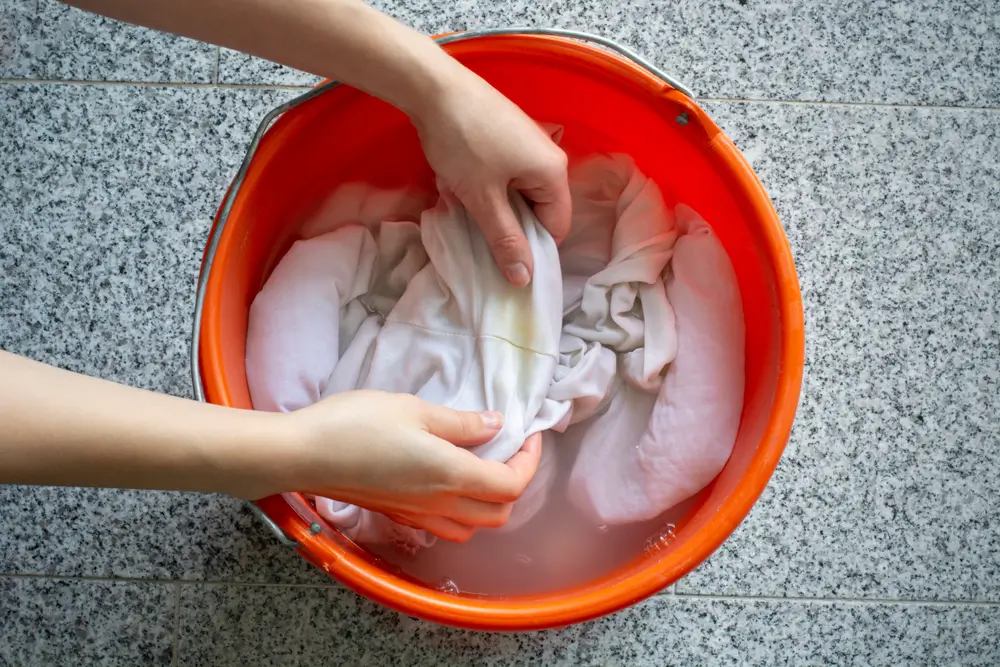 Women's hands washing dirty white shirts with yellow sweat stains to soak it in a bucket. 