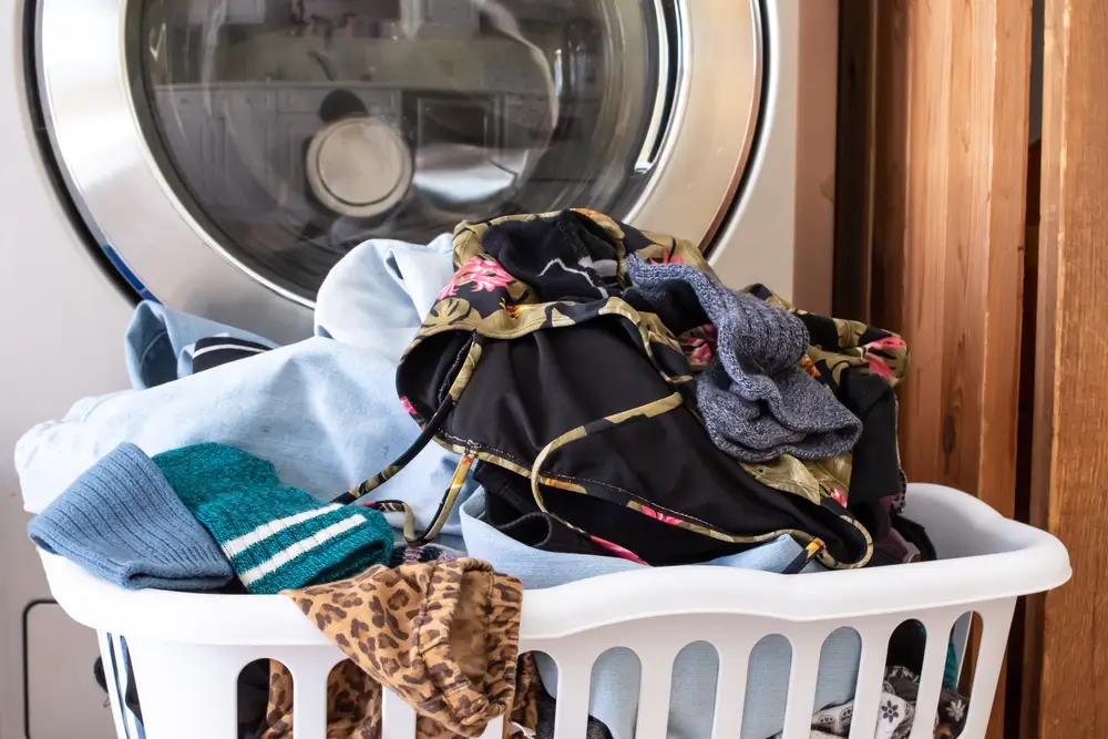 A pile of laundry sits in a square plastic hamper basket in front of a chrome style washing machine