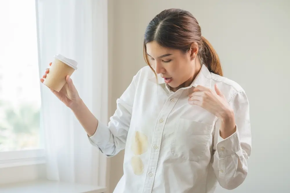 Cloth stain, disappointment asian young woman clumsy with hot coffee, tea stains on shirt, hand show making spill drop on white t-shirt, spot dirty or smudge on clothes at home, isolated on background
