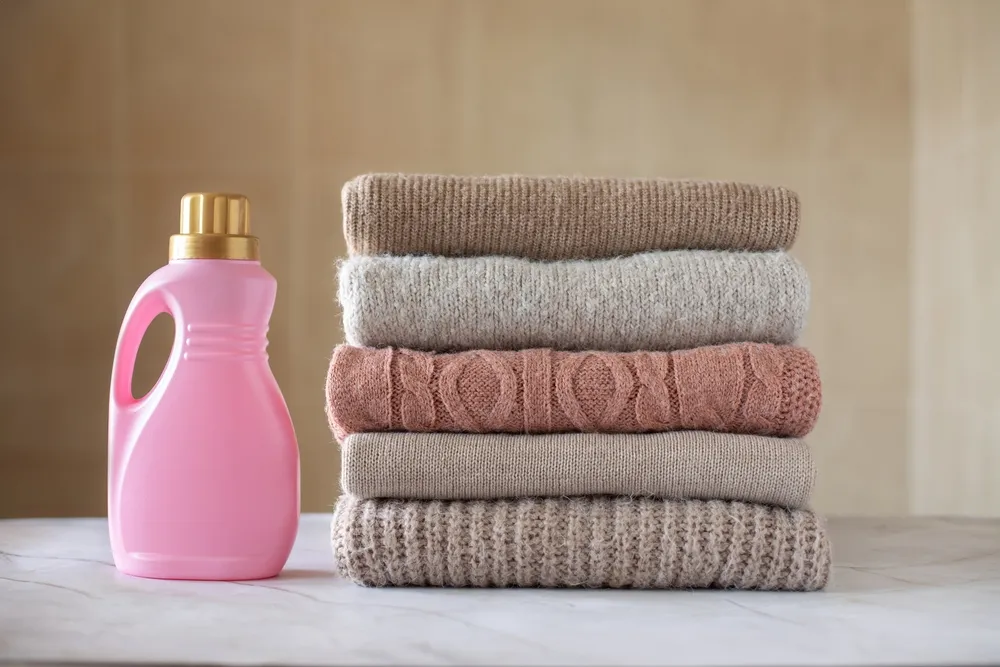 Bottle of detergent, fabric softener and a pile of clean colored sweaters on a marble table
