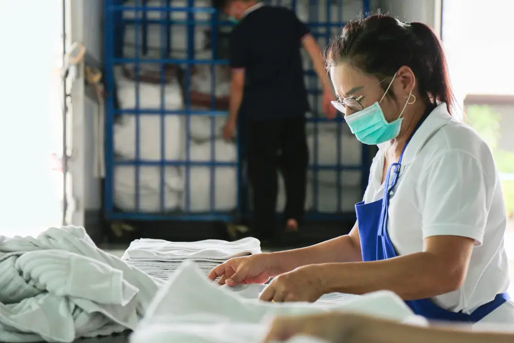 A laundry staff wearing a mask is folding towels with a blurred delivery truck in a background.