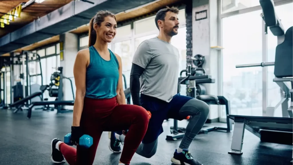 Athletic couple using dumbbells while exercising in lunge position in a gym.
