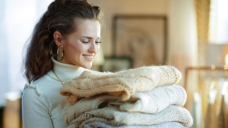 smiling stylish woman in white sweater and skirt in the modern house in sunny winter day looking at pile of sweaters.