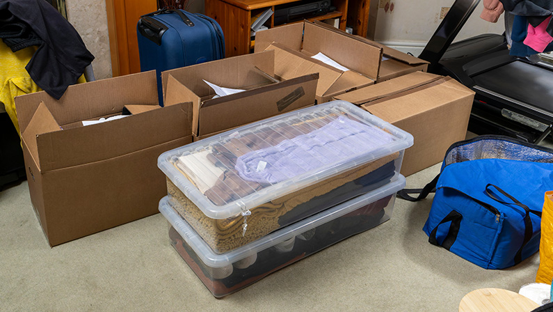 Clothes and belongings packed in cardboard boxes, luggage, and storage bins scattered around a living room