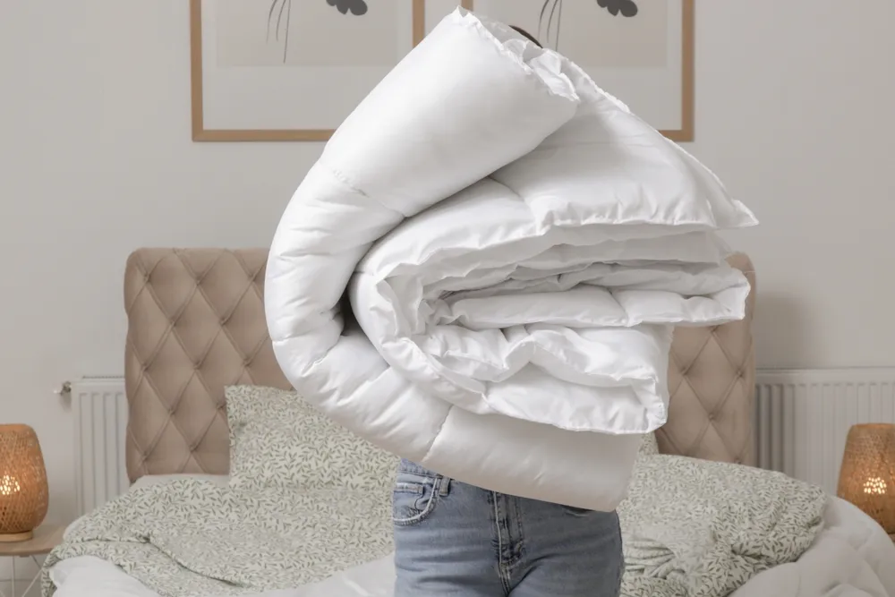 Woman holding pile of soft white folded duvet and pillows at home in her bedroom, cozy domestic lifestyle