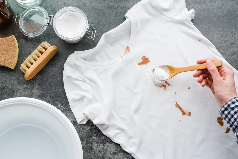 Hand applying baking soda powder to a white shirt with a visible barbeque stain, preparing for stain removal at home.
