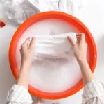 closeup of person hand washing clothes in an orange bucket