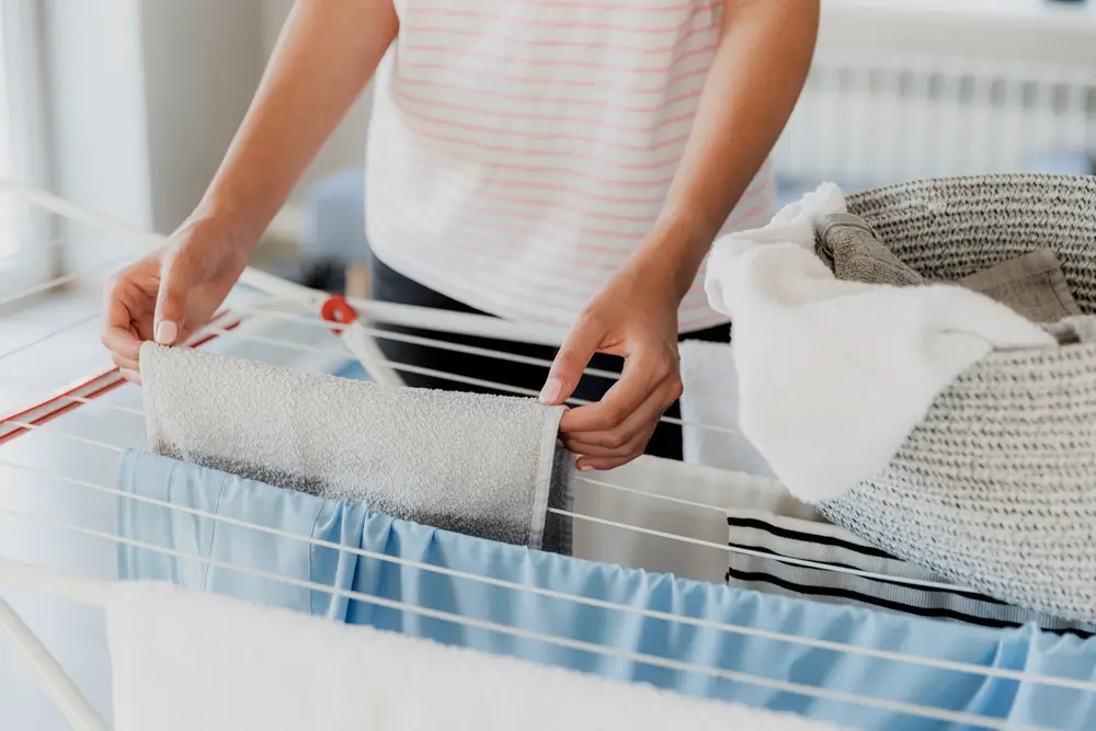 Woman hanging clean wet clothes laundry on drying rack at home laundry room.