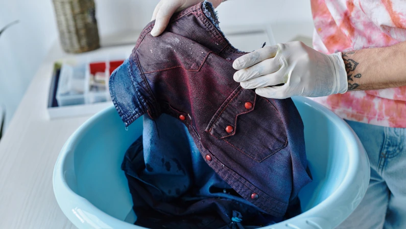 A young man in a tie-dye shirt washes a denim shirt in a blue plastic basin, reviving discarded clothing through sustainable DIY methods.