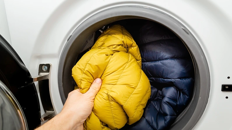 Putting winter jacket into the drum of open washing machine in laundry room. Washing dirty down jacket in the washer