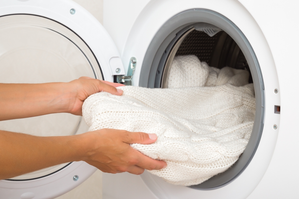 Young adult woman hands holding and unloading white warm soft sweater from opened washing machine