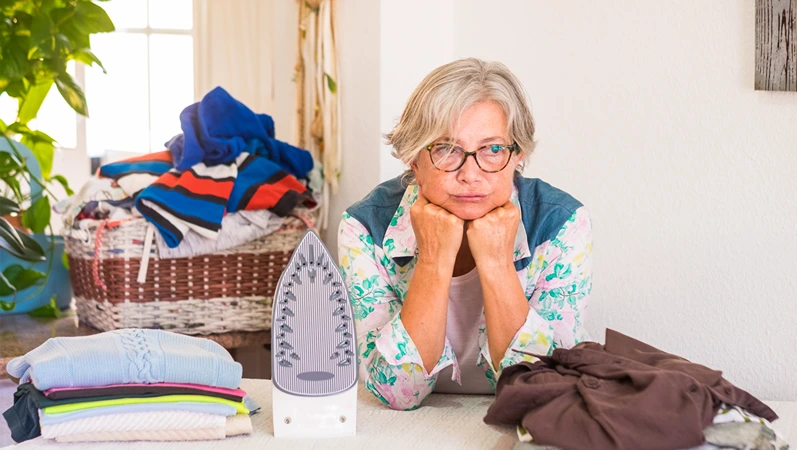 A senior woman with grey hair sad because she has too much clothes to iron. Corner of the domestic room with plants and white wall. One people only
