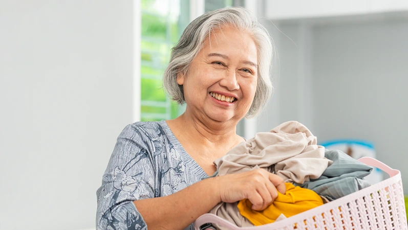 Smiling senior woman sorting laundry at home, Cheerful Asian woman holding a laundry basket filled with clothes