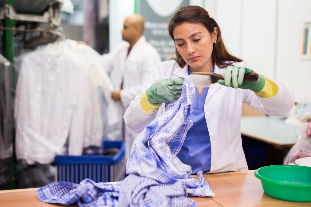 Focused female drycleaner removing spots and stains cleaning shirt using chemicals and brush