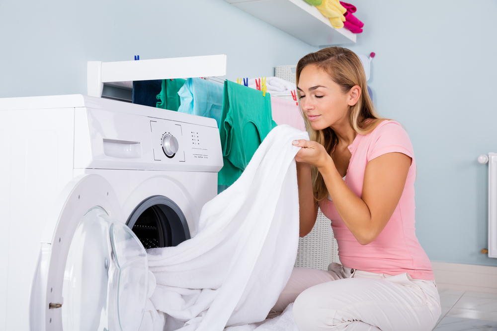 Young Woman Smelling Clean Clothes Near The Electronic Washer At Laundry Room