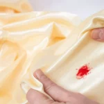 Woman hands stretching golden satin fabric with a red blood stain, soft focus close up