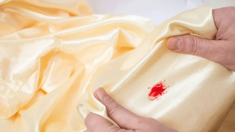 Woman hands stretching golden satin fabric with a red blood stain, soft focus close up