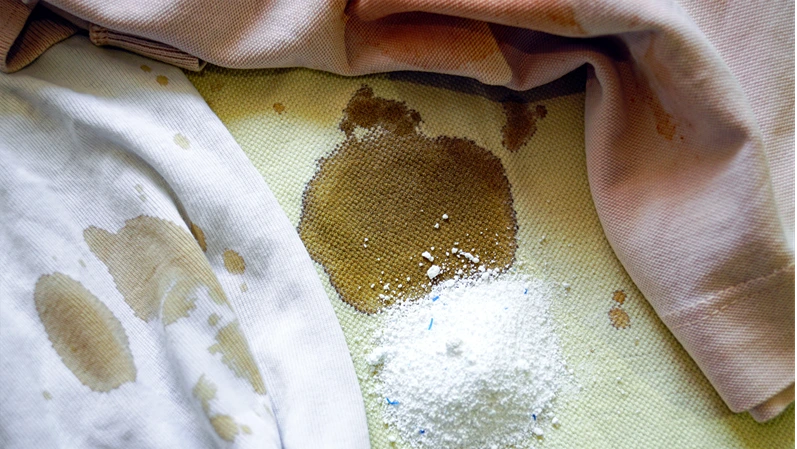 Removing stains from light-colored fabric using washing powder: photo of stained cotton T-shirts in white, beige, and light pink with a handful of washing powder poured on them.
