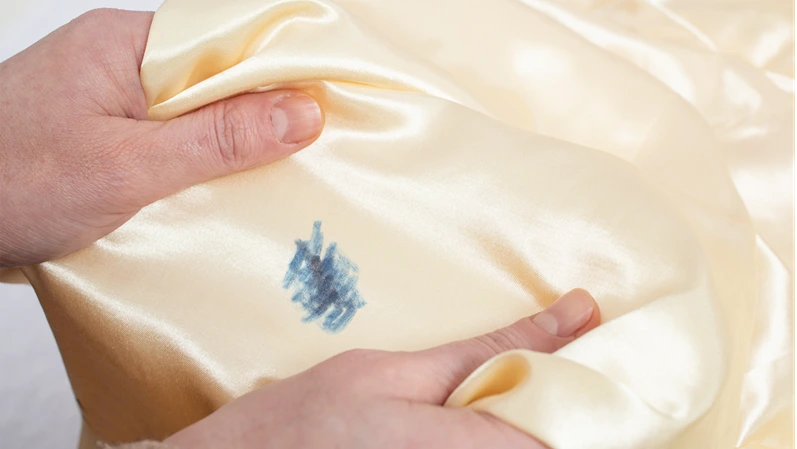Woman hands stretching golden satin fabric with a blue ink stain, soft focus close up