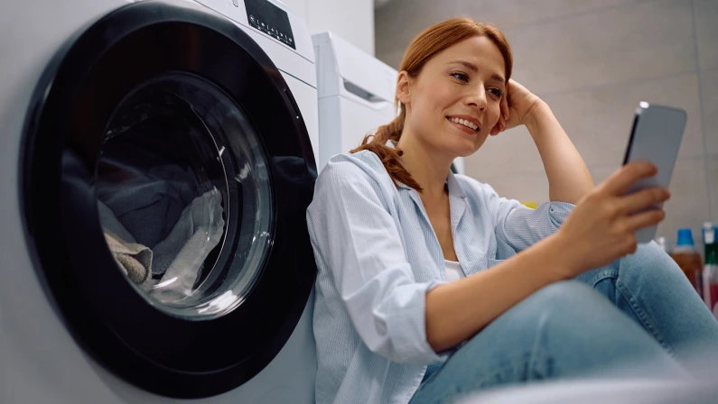 Happy woman using mobile phone while waiting for washing machine to finish in laundry room. Copy space.